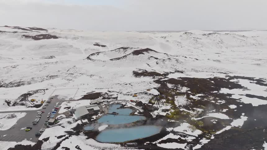 A stunning aerial pullback reveals a geothermal spa with steaming blue pools nestled in the vast, snow-covered volcanic landscape of Iceland