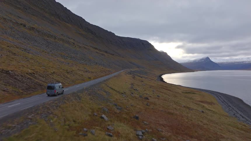 A campervan drives along a picturesque Icelandic fjord road, nestled between a rugged mountain and the sea