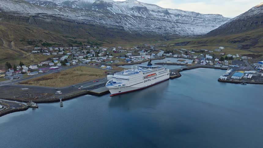 A slow aerial pullback reveals the Smyril Line ferry docked in a tranquil Icelandic harbor, surrounded by a small town and majestic, snow-capped mountains