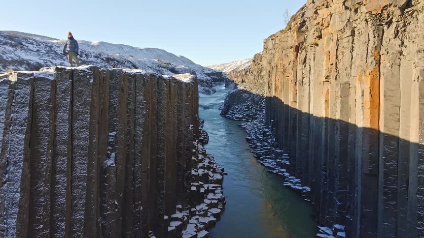 A lone hiker stands atop the spectacular snowy basalt columns of Studlagil Canyon in Iceland, revealed by a dramatic aerial pullback shot