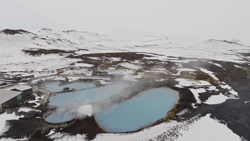 A slow orbiting aerial shot reveals a stunning geothermal spa with steaming, milky blue pools contrasting with the surrounding snowy volcanic landscape