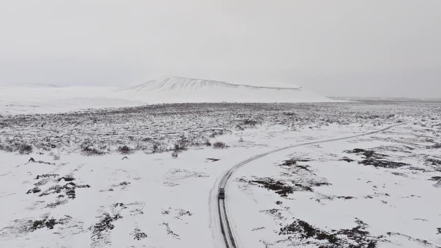 An aerial view follows a camper van driving on a remote road through a vast, snow-covered volcanic landscape towards a large crater in Iceland