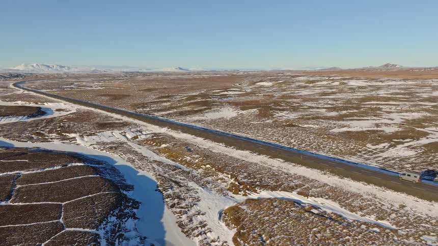 An aerial view follows a lone camper van driving along a remote highway through a vast, snow-dusted landscape on a clear winter day