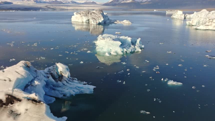 A cinematic aerial shot pulls back from a large iceberg to reveal the majestic Jökulsárlón glacier lagoon in Iceland, dotted with ice and surrounded by snow-covered mountains