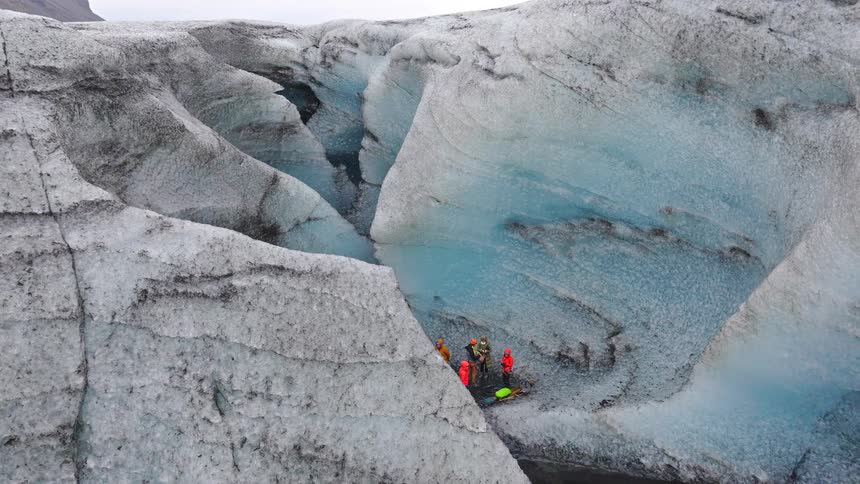 An aerial shot pulls back from the deep blue ice of a glacier to reveal a group of hikers exploring its massive crevasses