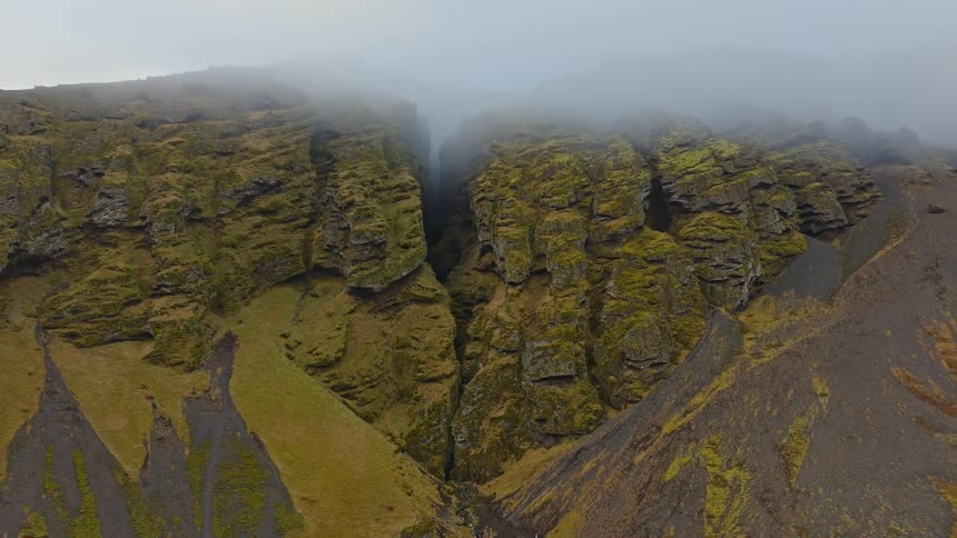 A moody aerial view pulls back from a rugged, mossy cliff to reveal a deep gorge with a hidden waterfall shrouded in thick mist