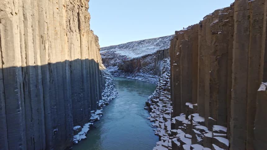 Aerial drone slowly flies through a majestic, snow-covered basalt column canyon with a river flowing below in Iceland