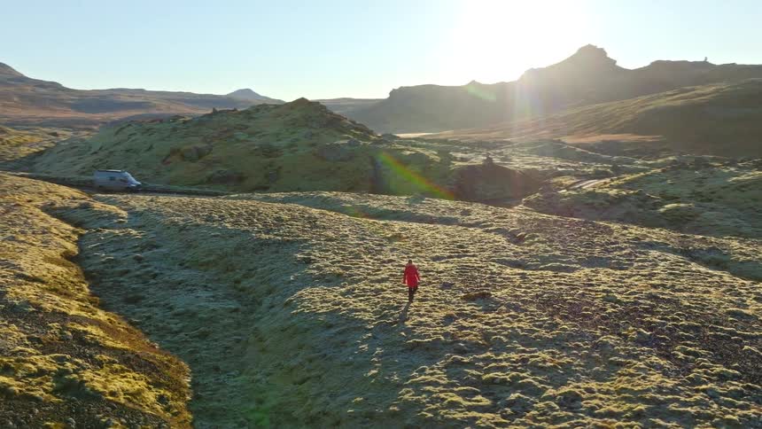 An aerial view follows a solo adventurer in a red coat hiking across the vast, moss-covered volcanic landscape of Iceland at sunrise