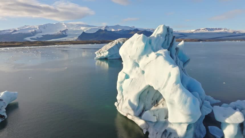 A serene aerial shot slowly pans around a majestic, sculpted iceberg floating in a tranquil glacial lagoon with snow-covered mountains in the background
