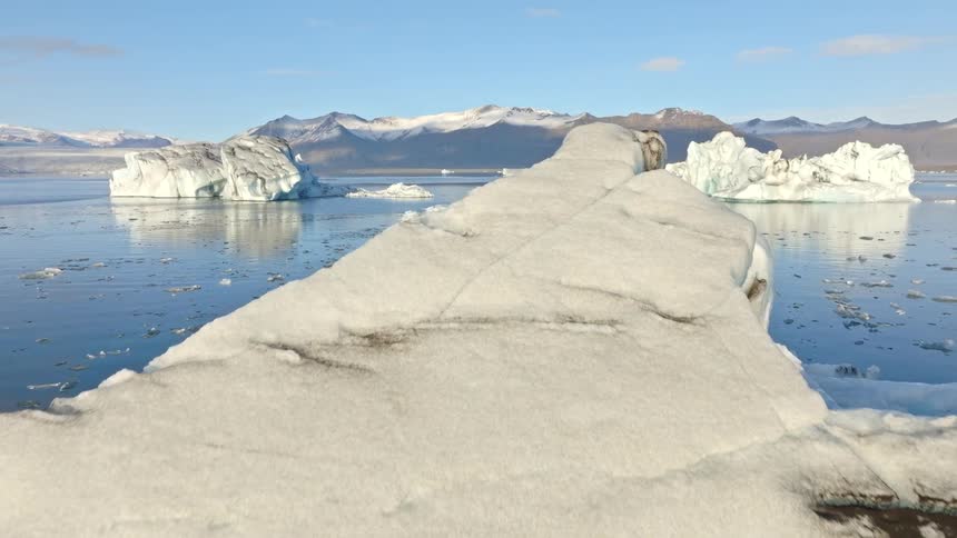 Aerial drone footage flies over a massive iceberg, revealing a beautiful glacial lagoon filled with ice formations against a mountain backdrop