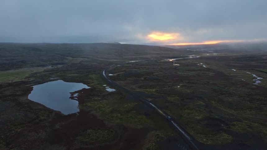 An aerial perspective follows a single car traveling on a remote dirt road through a vast, moody Icelandic landscape at twilight
