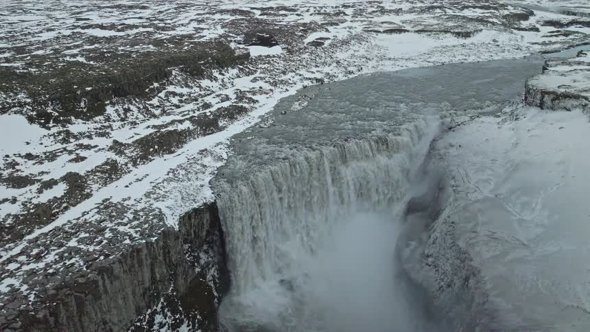 Aerial footage pulls back to reveal a massive, powerful waterfall cascading through a vast, snow-covered canyon in winter