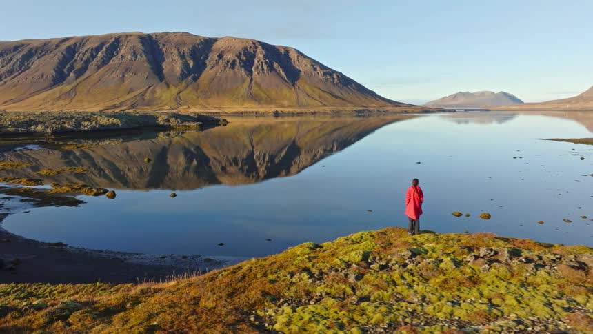Cinematic aerial shot of a person in a red coat standing on a mossy hill, looking out at a stunning mountain reflection in a still lake at sunrise