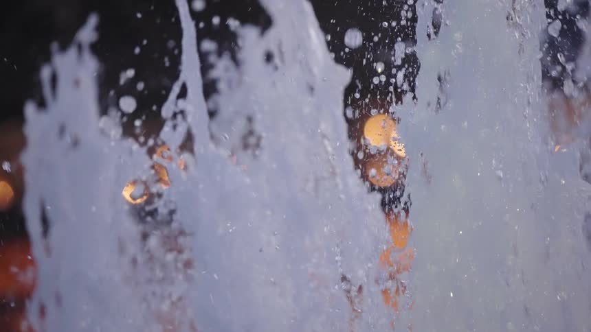 Bubbling water from a fountain splashes in mesmerizing slow motion against a backdrop of glowing bokeh city lights at night