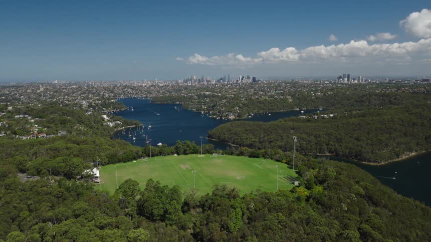 Static aerial shot of a green sports field surrounded by lush forest, overlooking a beautiful bay and a major city skyline on a sunny day