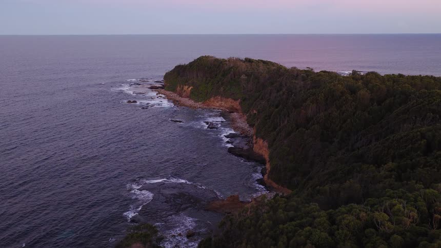 A serene aerial shot captures a forested headland on a rocky coast as gentle waves break at dusk
