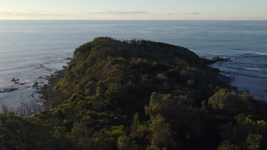 A majestic aerial view shows a lush green headland bathed in the golden light of sunrise, extending into the calm, vast ocean
