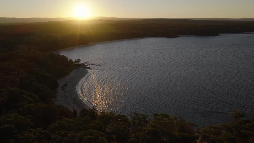 A serene aerial view shows the sun setting over a forested coastline, casting a golden reflection on the calm waters of a secluded bay
