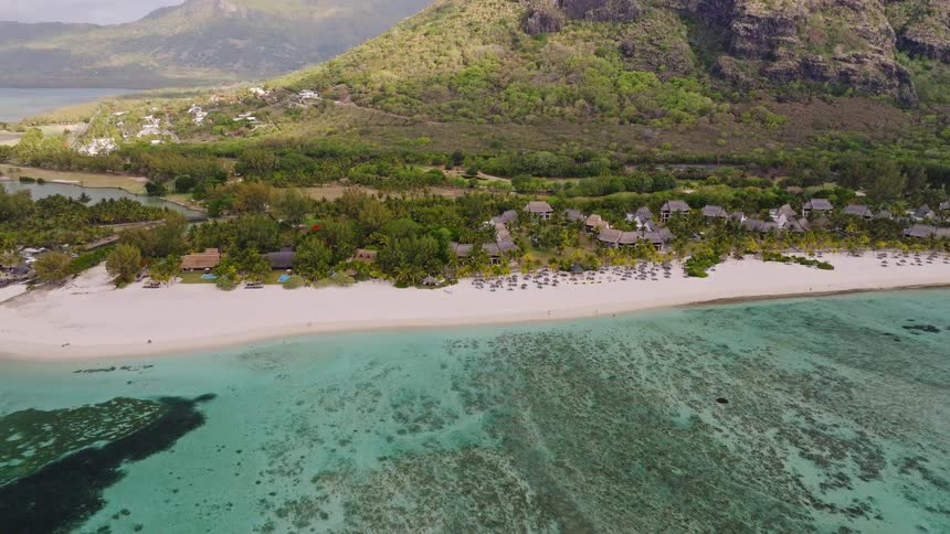 An aerial view reveals a stunning tropical paradise, showcasing a white sand beach and luxury resort nestled at the foot of an iconic mountain
