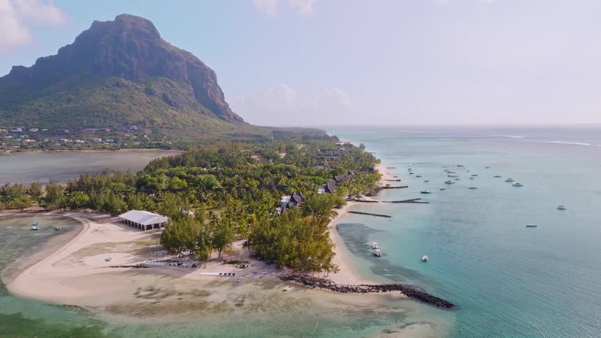 Stunning aerial reveal shot of a tropical beach on the coast of Mauritius, showcasing the crystal clear turquoise ocean, anchored boats, and the magnificent Le Morne Brabant mountain
