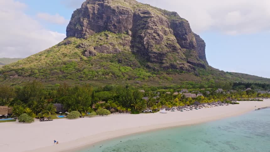 Aerial view pulls back from a wedding gazebo on a white sand beach to reveal a luxury tropical resort at the base of an iconic mountain in Mauritius