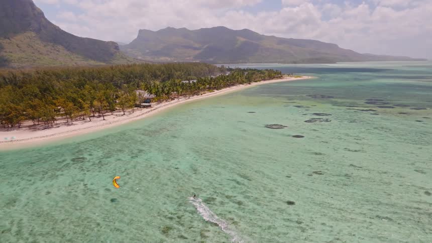 An aerial view captures a kitesurfer gliding across the stunning turquoise lagoon of a tropical island with a majestic mountain in the background