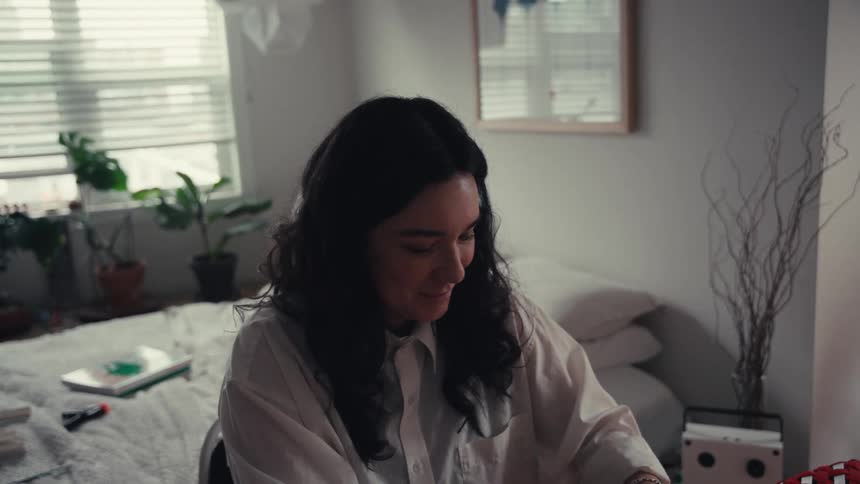 A young woman with dark curly hair smiles while concentrating on a project in her peaceful, plant-filled bedroom