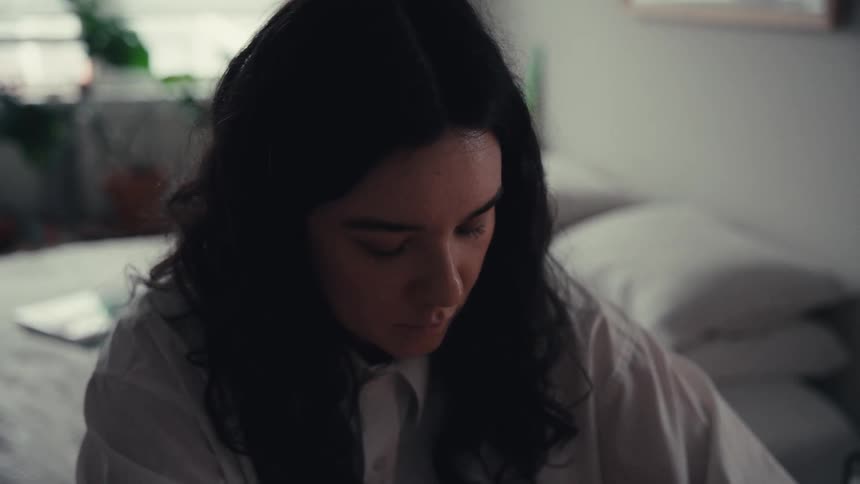 A young woman with dark hair sits in her dimly lit bedroom, deeply focused on her work