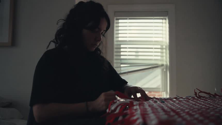 A focused woman with dark hair carefully weaves a red and white patterned craft in a softly lit room