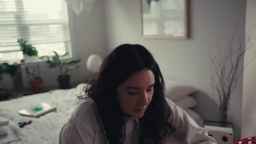 A young woman with dark hair focuses on her creative project in a naturally lit bedroom