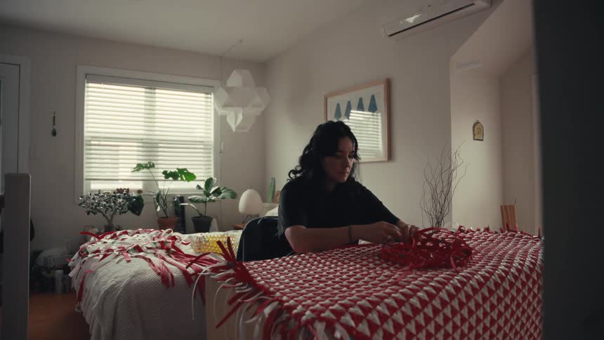 An artist meticulously weaves an intricate red and white patterned tapestry in her bright and airy apartment