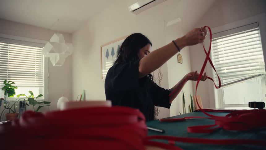 A focused woman carefully unravels a long red strap in slow motion while working at her desk in a sunlit home studio