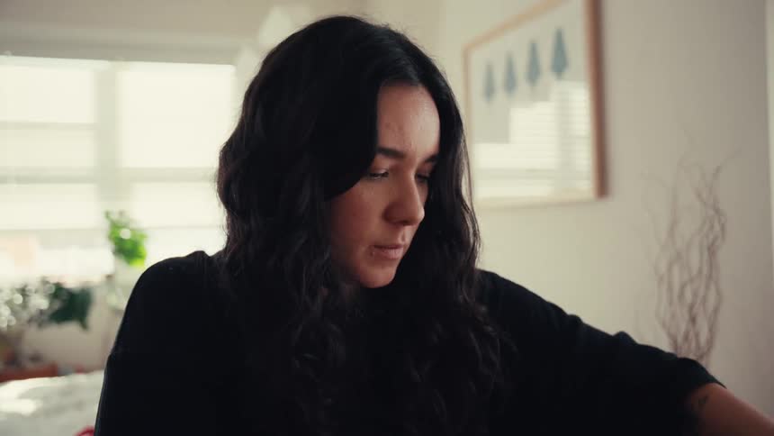 A young woman with long dark hair thoughtfully prepares a gift with a red ribbon in her naturally lit apartment