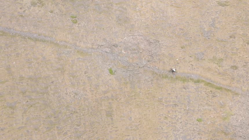 An aerial top-down view shows a lone mountain biker navigating a rugged, rocky trail through a vast, arid landscape