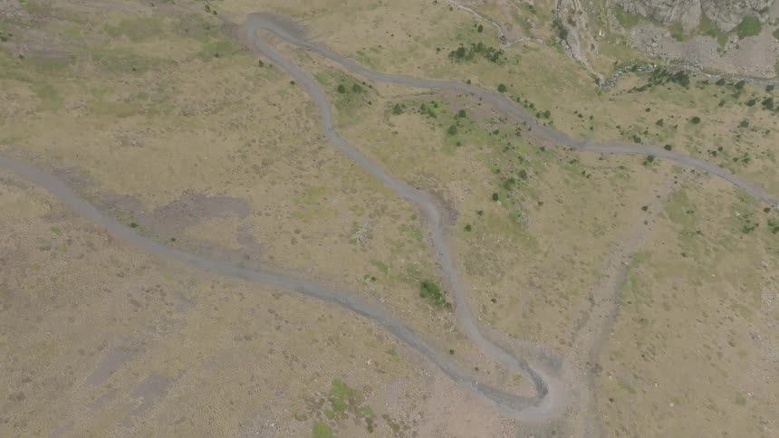 A static bird's-eye aerial view shows a winding dirt road with hairpin turns traversing a grassy mountain valley
