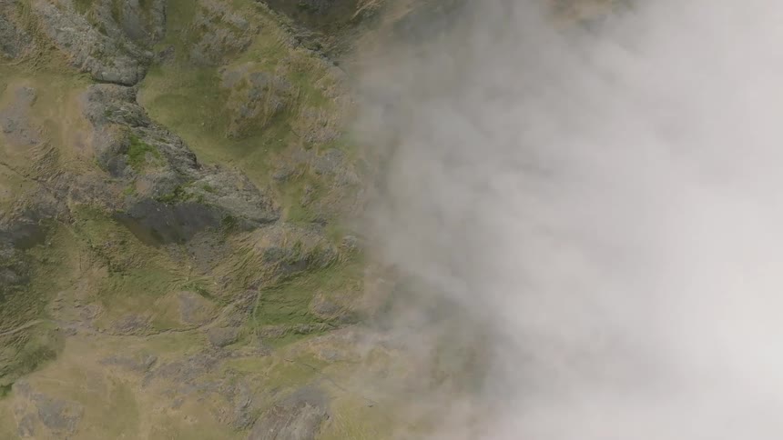 An aerial top-down view shows thick white clouds slowly moving across a rugged, green mountain landscape