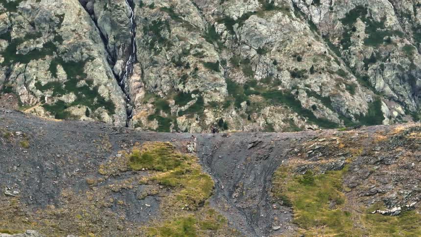 Two distant hikers trek across a vast, rugged mountain landscape with a small waterfall in the background