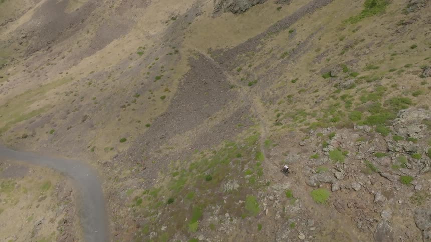 A dramatic aerial shot tilts down from a cloudy mountain range to a lone hiker trekking along a steep, winding trail