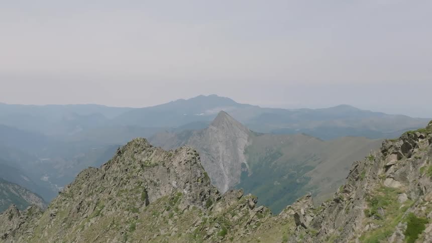 An aerial view slowly advances over a green and rocky mountain ridge, revealing a vast and hazy mountain range in the distance
