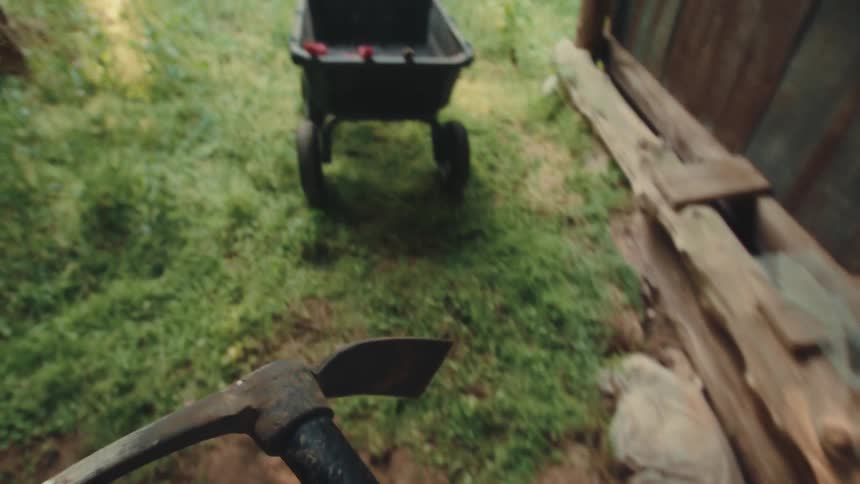 From a first-person perspective, a person grabs a pickaxe from a dark, rustic shed and walks outside into a grassy yard