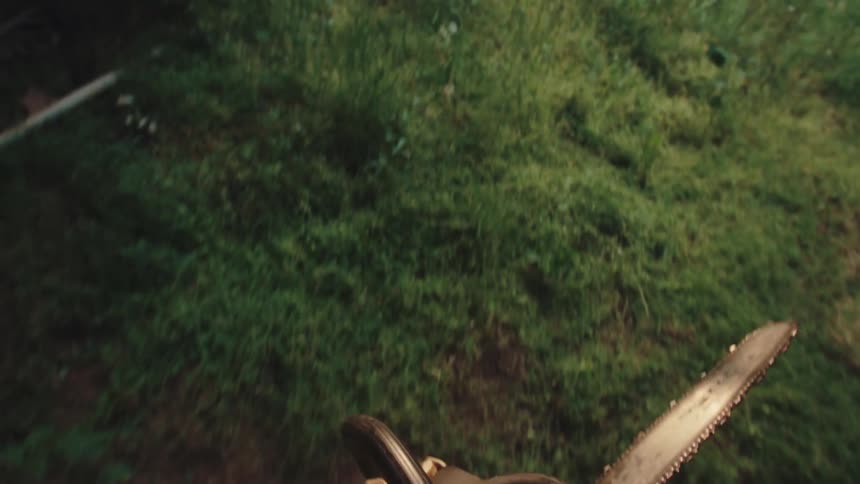 A person's first-person perspective of picking up and starting a chainsaw in the evening before walking through an overgrown field