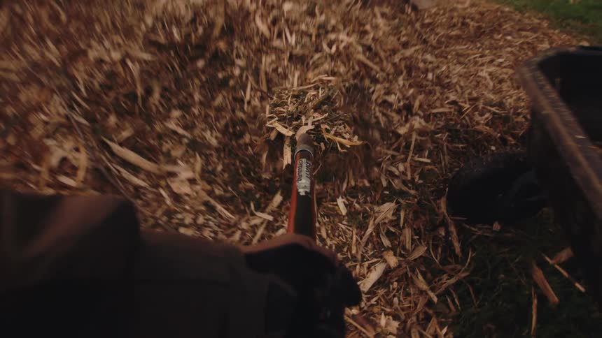 A first-person view of shoveling wood chip mulch from a large pile into a wheelbarrow for yard work