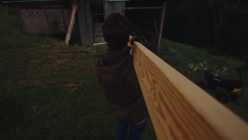 POV shot from a wooden plank as a young man carries it towards a rustic barn in slow motion at dusk