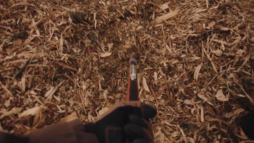 A first-person view of a gardener wearing gloves and using a shovel to scoop up wood chip mulch into a wheelbarrow