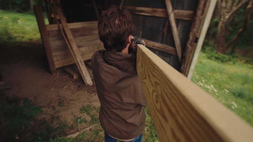 A man wearing work gloves carries a long wooden plank out of a dark barn and into a grassy backyard during golden hour