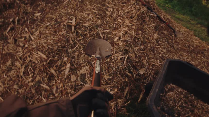A dizzying first-person perspective of shoveling mulch from a large pile into a wheelbarrow during yard work