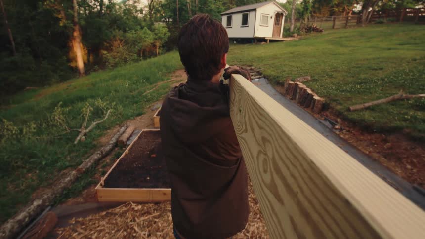 Slow motion following shot of a person carrying a heavy wooden plank across a yard to build raised garden beds at dusk