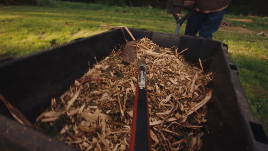 First-person perspective of a shovel riding in a wheelbarrow full of wood chips being transported across a lawn
