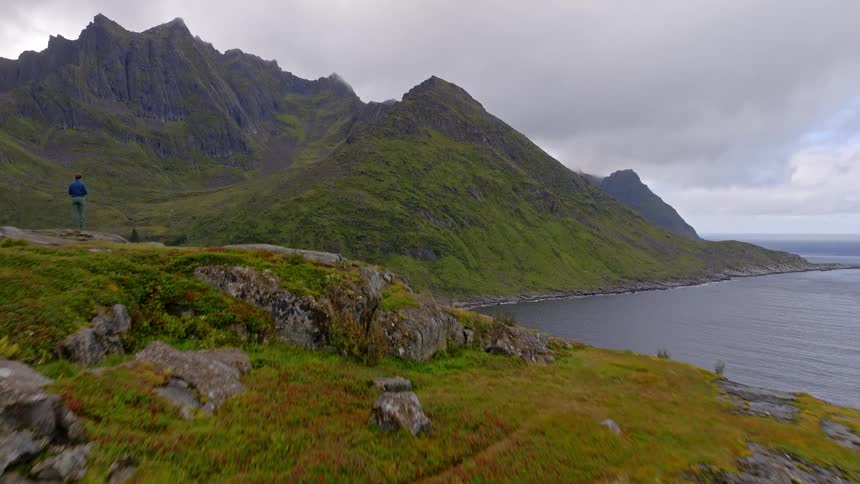 An aerial shot flies over a hiker standing on a grassy cliff overlooking a dramatic mountainous coastline in Norway
