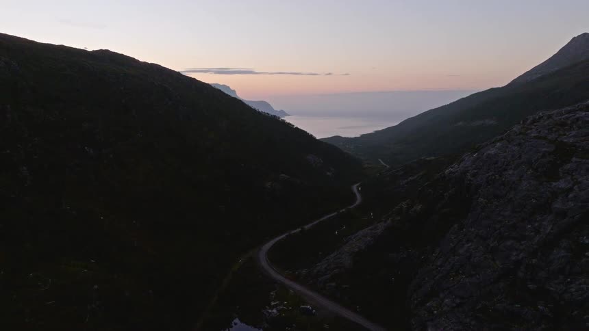 A serene aerial view slowly pulls back over a winding road in a dark mountain valley that leads to the ocean at dusk